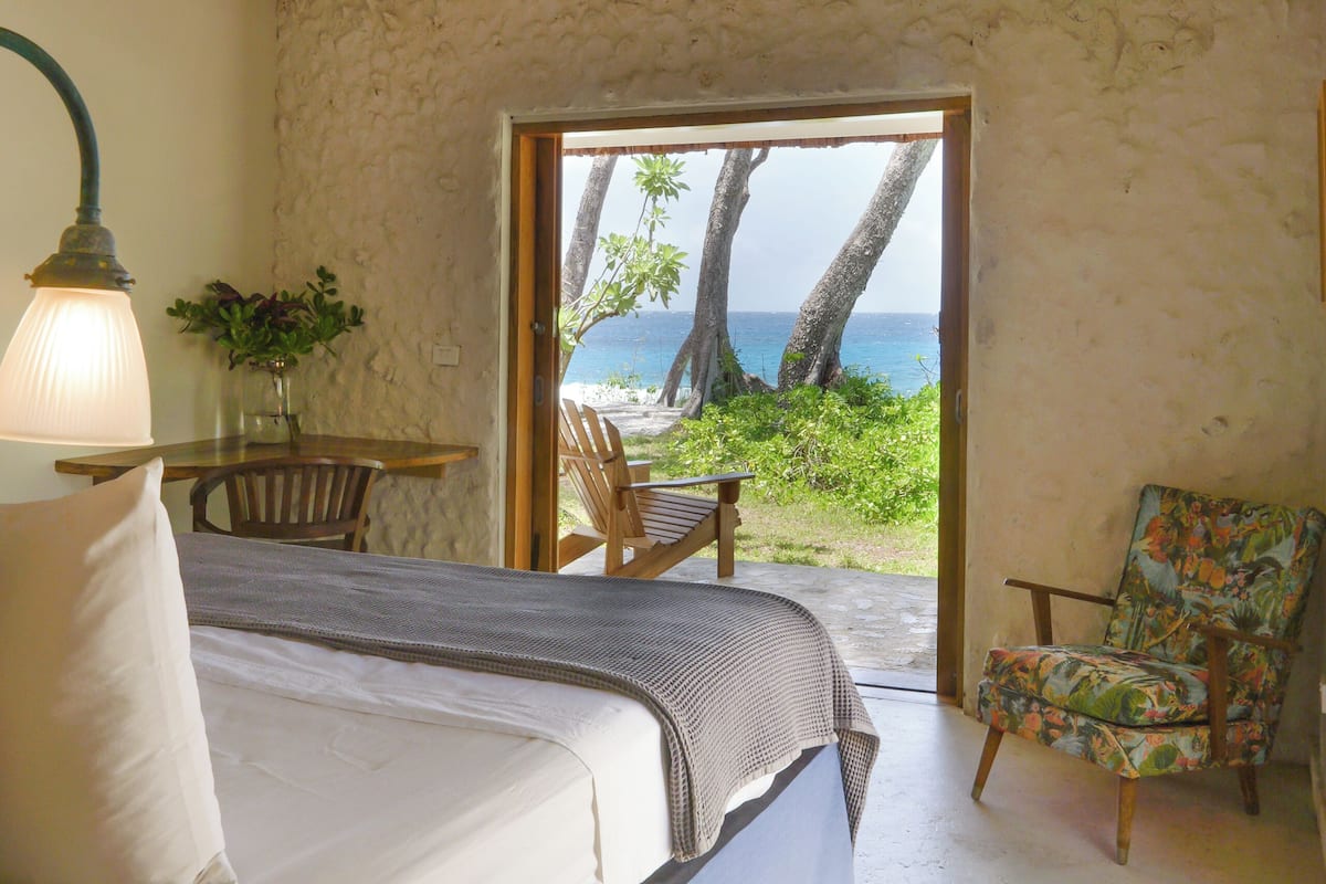 Quarto com cama, uma cadeira, uma mesa e vista para a praia.