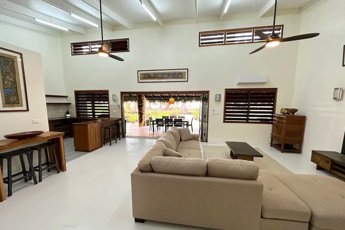 Uma sala de estar ampla com um sofá seccional bege grande, uma cozinha com banquetas de madeira e uma área de jantar com mesa e cadeiras.