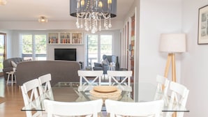 Un salon moderne avec une table à manger en verre, des chaises blanches, un canapé gris, un lustre et une bibliothèque.