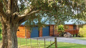 A wooden cabin with a large tree in front, a paved driveway, and a blue garage door. - Tranquility at Cascade (Norfolk Island)