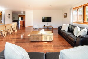 Ampla sala de estar com mesa de jantar, sofá, televisão e cozinha ao fundo.
