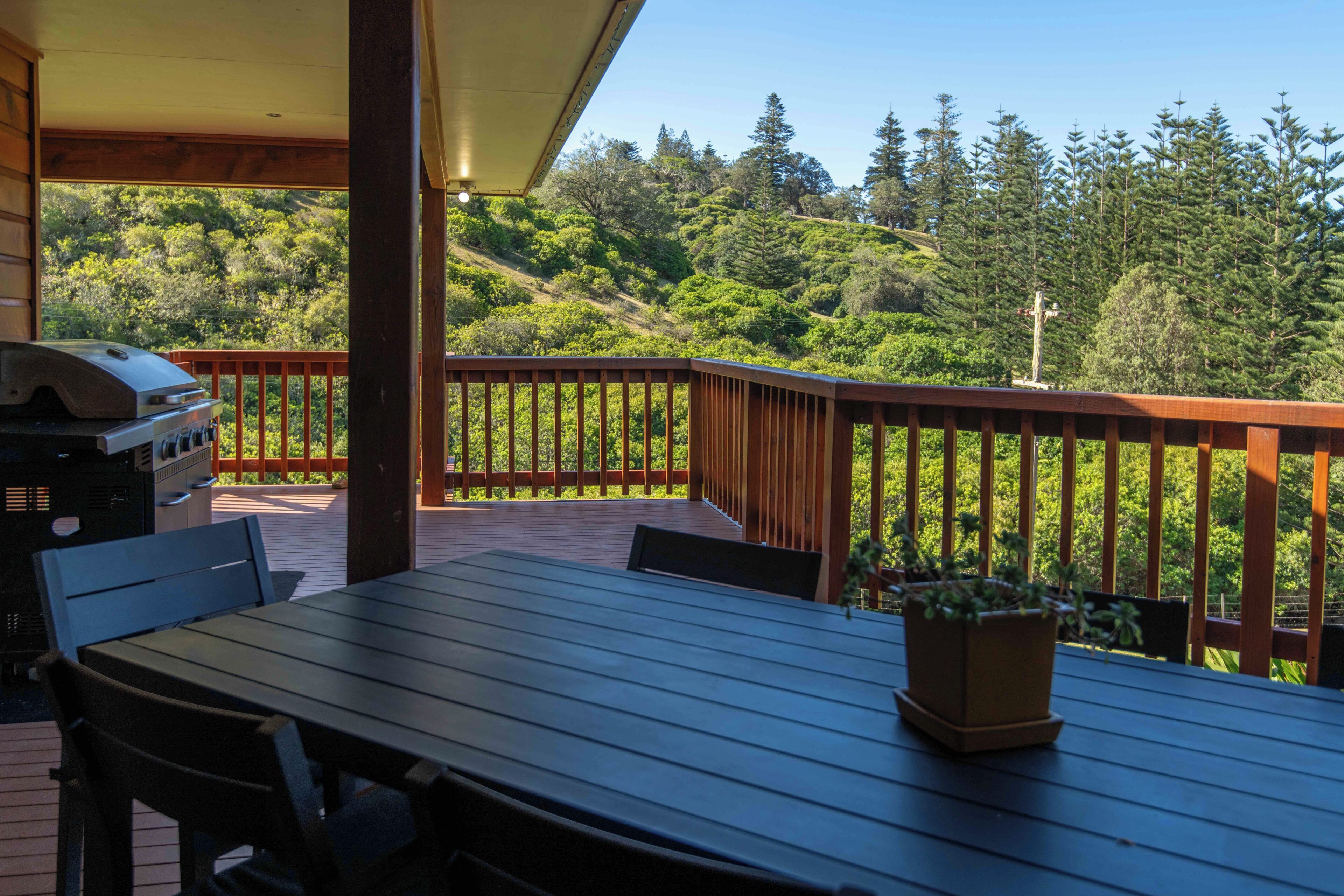 A wooden deck with a table and chairs, a grill, and a view of a forested hillside.