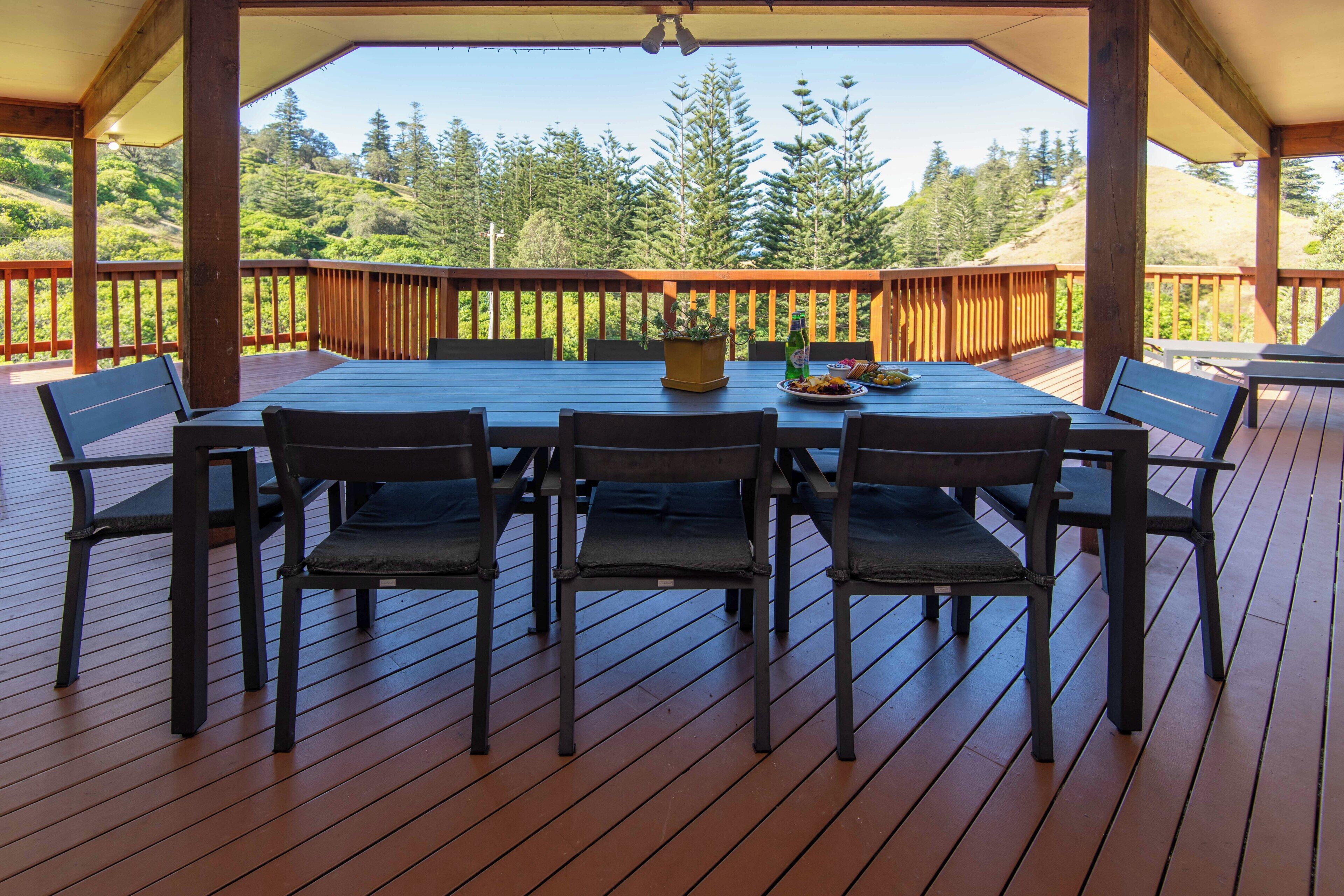 Terraza al aire libre con mesa y sillas de madera, rodeada por barandilla de madera, con vistas a un paisaje boscoso.