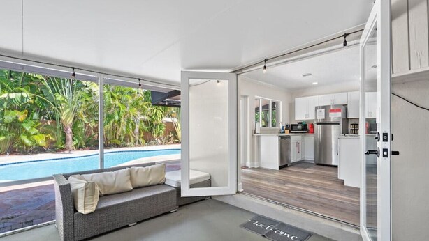 A modern kitchen with white cabinets and stainless steel appliances, a dining area with a gray sofa and a view of a swimming pool.