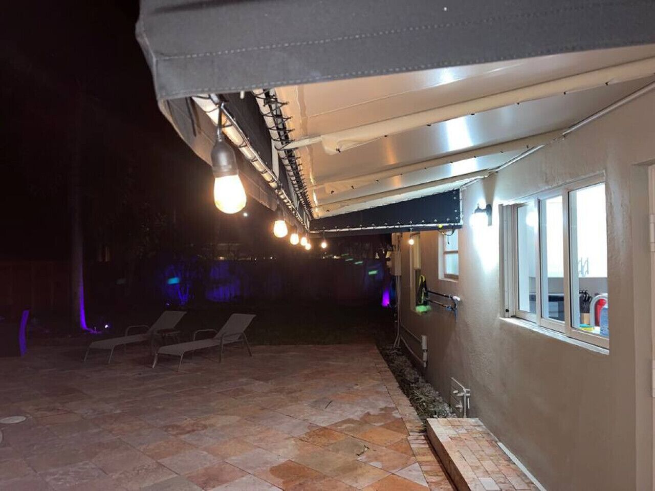 An outdoor patio area with stone flooring and lounge chairs, under a covered section with hanging lights.