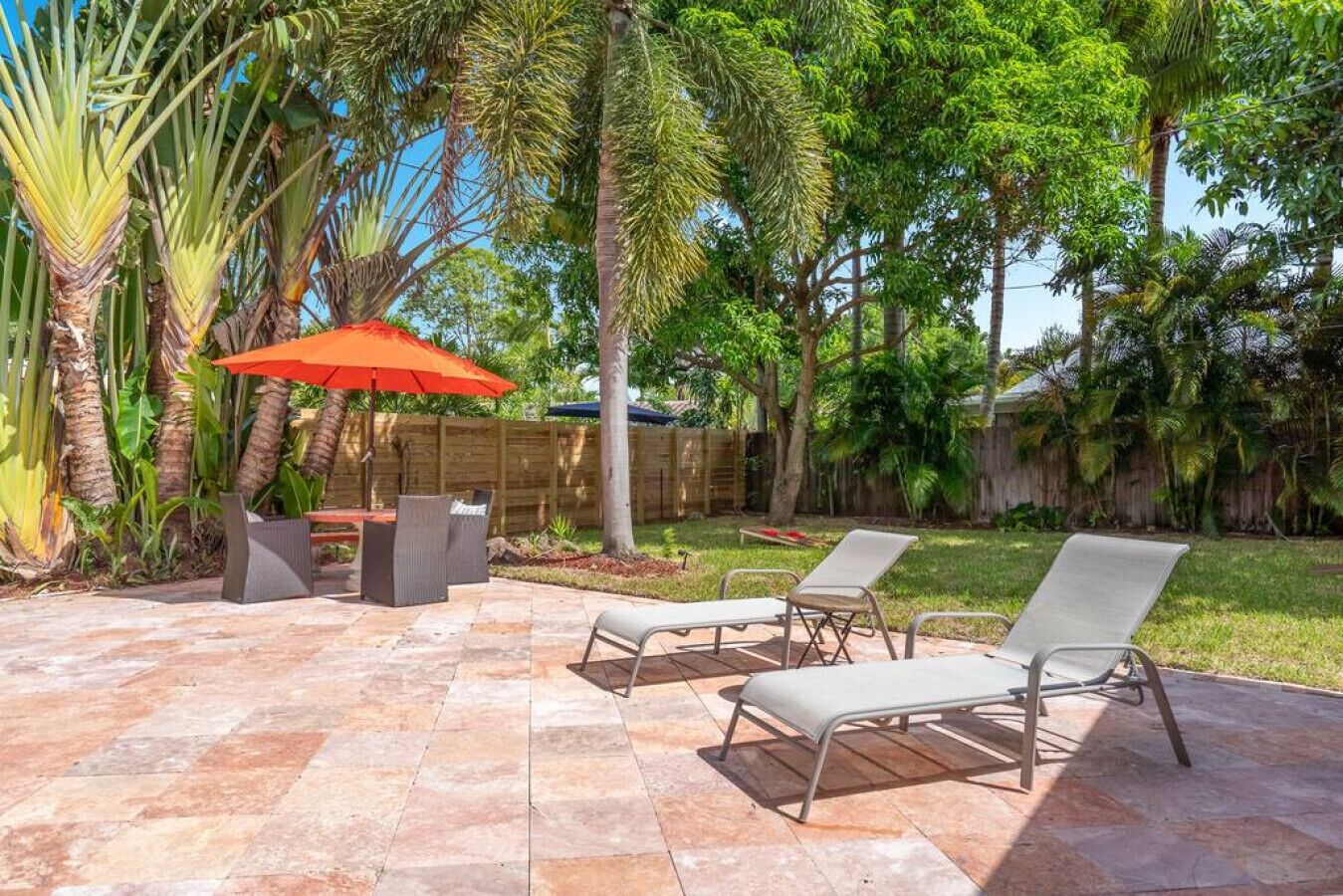 An outdoor patio with stone flooring, a wooden table, two lounge chairs, and a red umbrella.