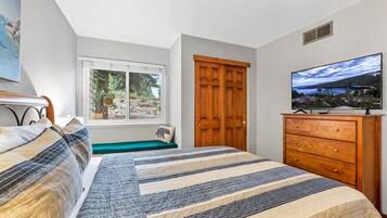 A bedroom with a bed, a wooden dresser, a television, a window with a view of greenery, and a door.