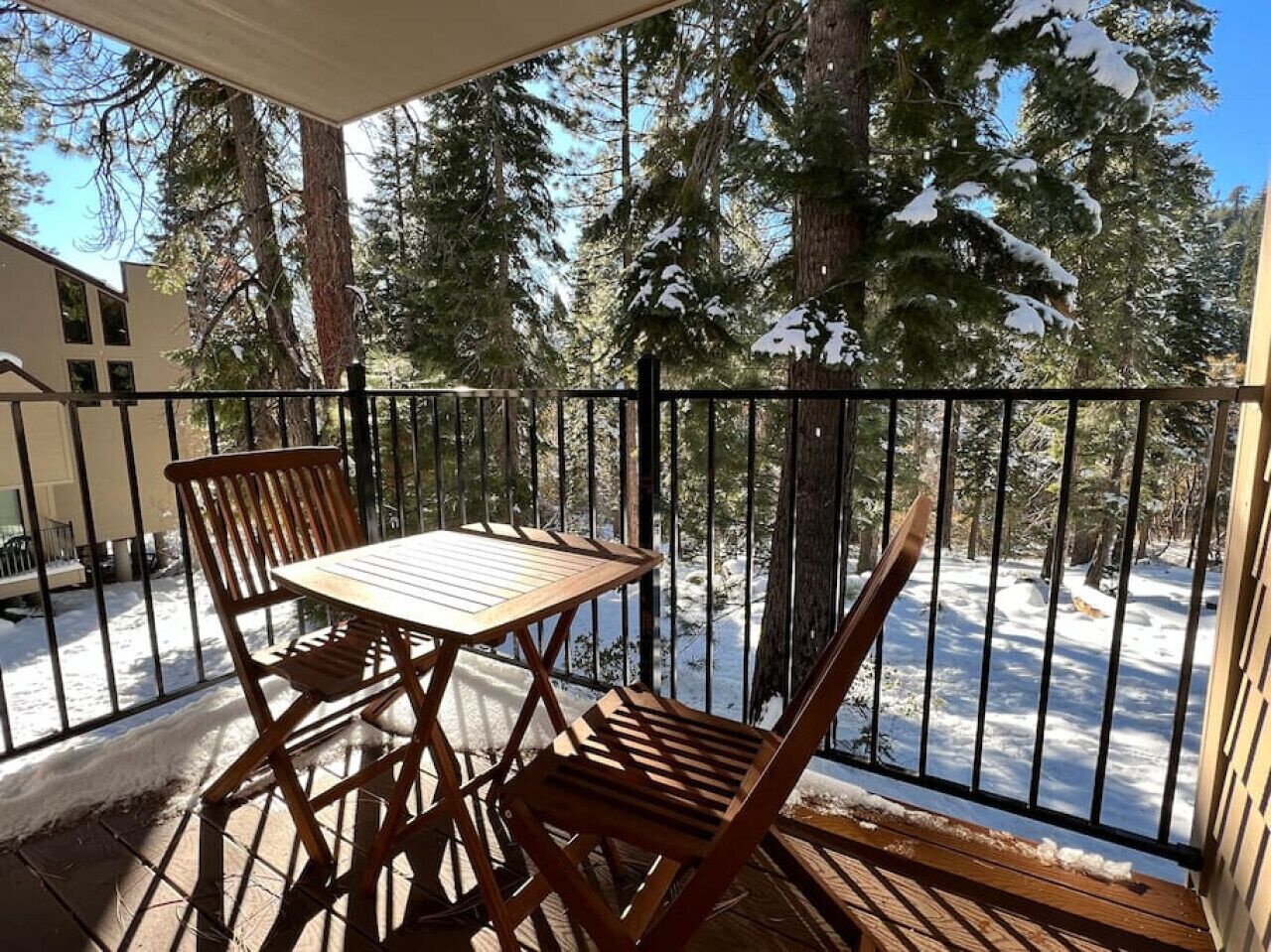A wooden deck with a table and chairs, surrounded by a metal railing, overlooking a snowy landscape with trees.