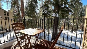 A wooden deck with a table and chairs, surrounded by a metal railing, overlooking a snowy landscape with trees.