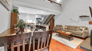 Ein geräumiger Wohnbereich mit Esstisch, Holzstühlen, einer Couch und einem Couchtisch. Im Hintergrund ist eine Küche mit weißen Schränken und einer Treppe zu einer oberen Ebene zu sehen.