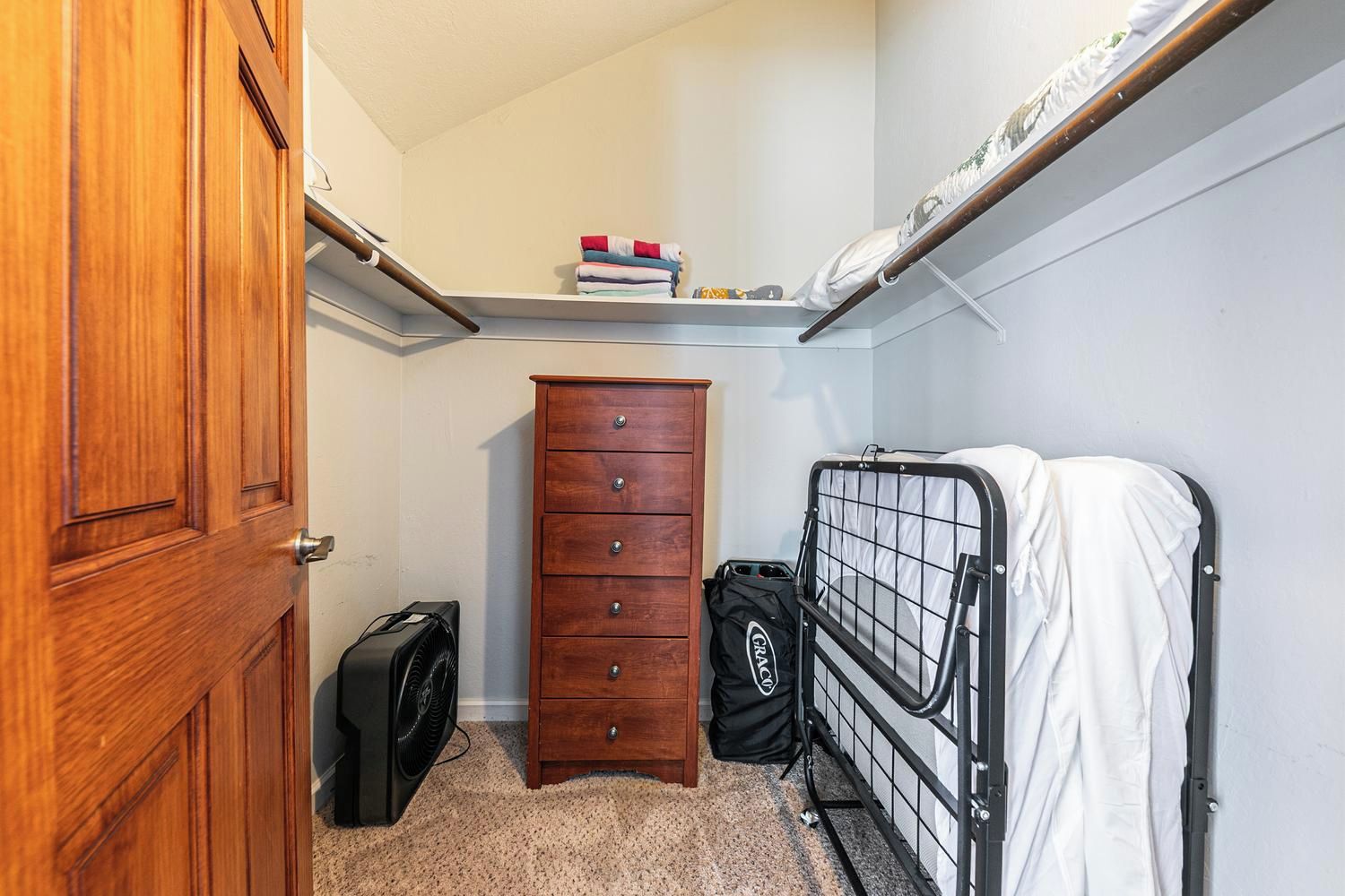 A compact room with a wooden door, a dresser, a fan, a bed with a metal frame, and a shelf with folded clothes.