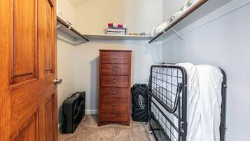 A compact room with a wooden door, a dresser, a fan, a bed with a metal frame, and a shelf with folded clothes.