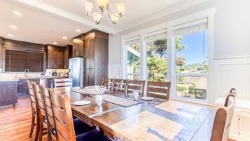 A spacious dining area with a large wooden table and chairs, a kitchen with dark cabinetry, and large windows offering a view of the outdoors.