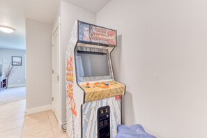 Una máquina de videojuegos retro con pantalla y panel de control, ubicada en una habitación con piso de baldosas y una puerta.