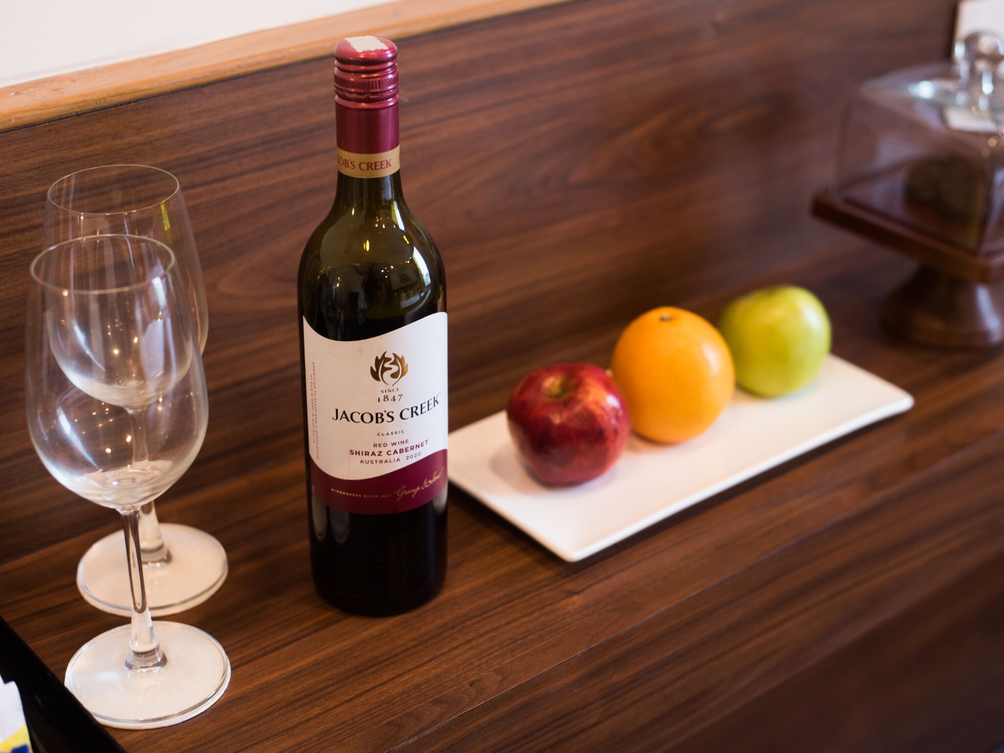 A bottle of Jacob's Creek wine, a wine glass, and a plate with an apple, an orange, and a green apple on a wooden table.