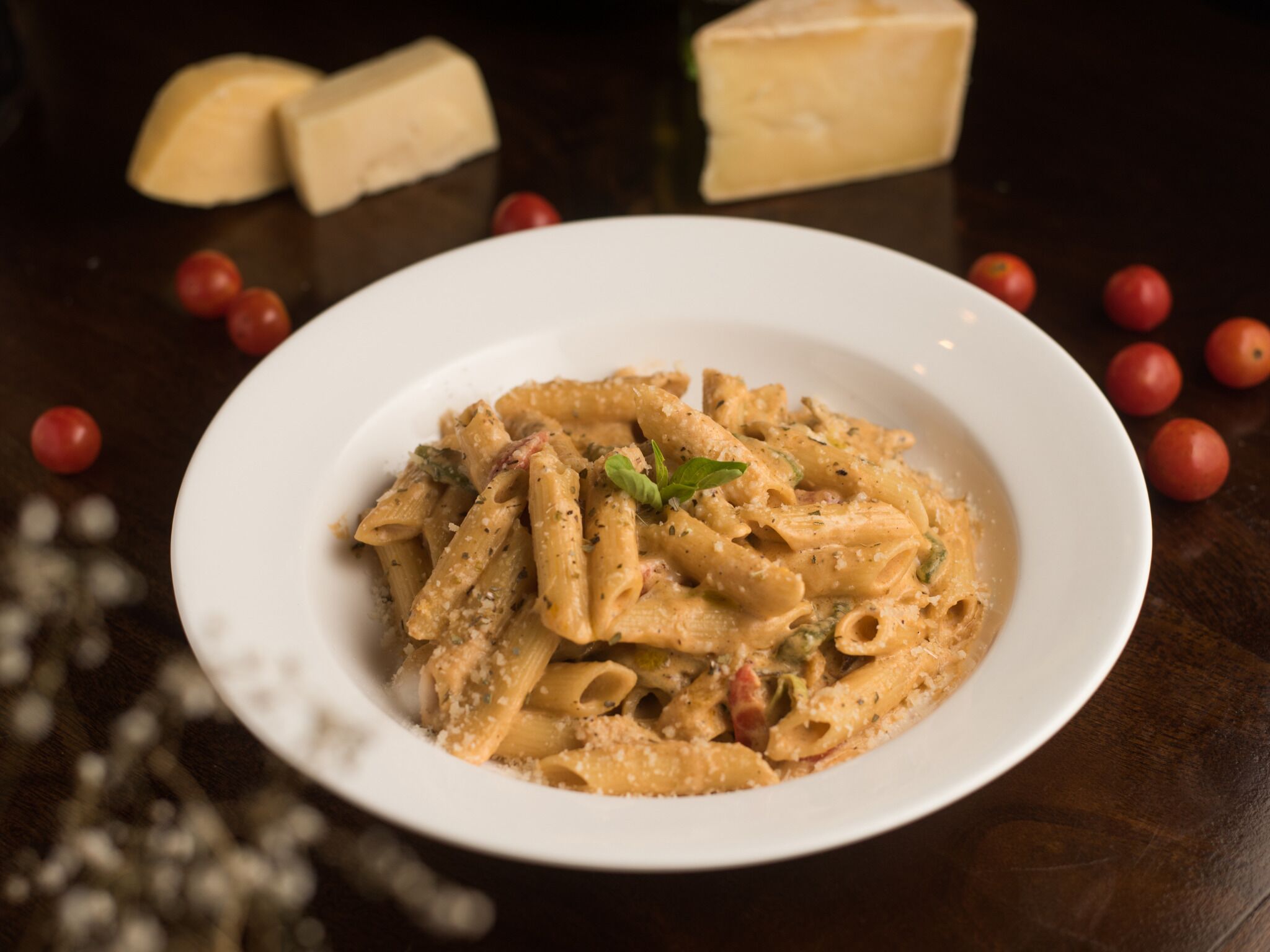 A white plate of pasta with cheese and herbs, surrounded by cherry tomatoes and cheese on a dark wooden table.