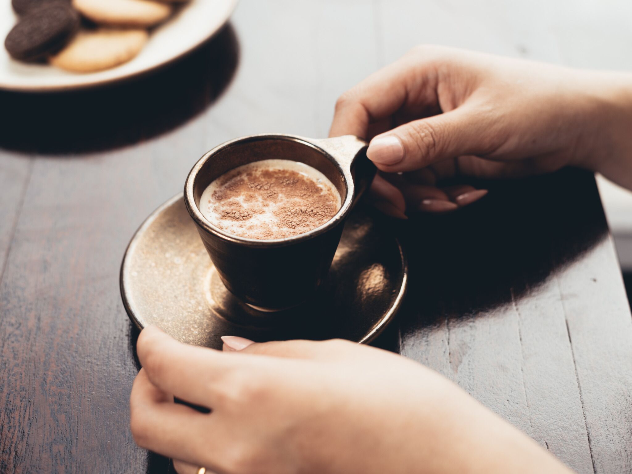 A cup of coffee on a saucer with a plate of cookies in the background.