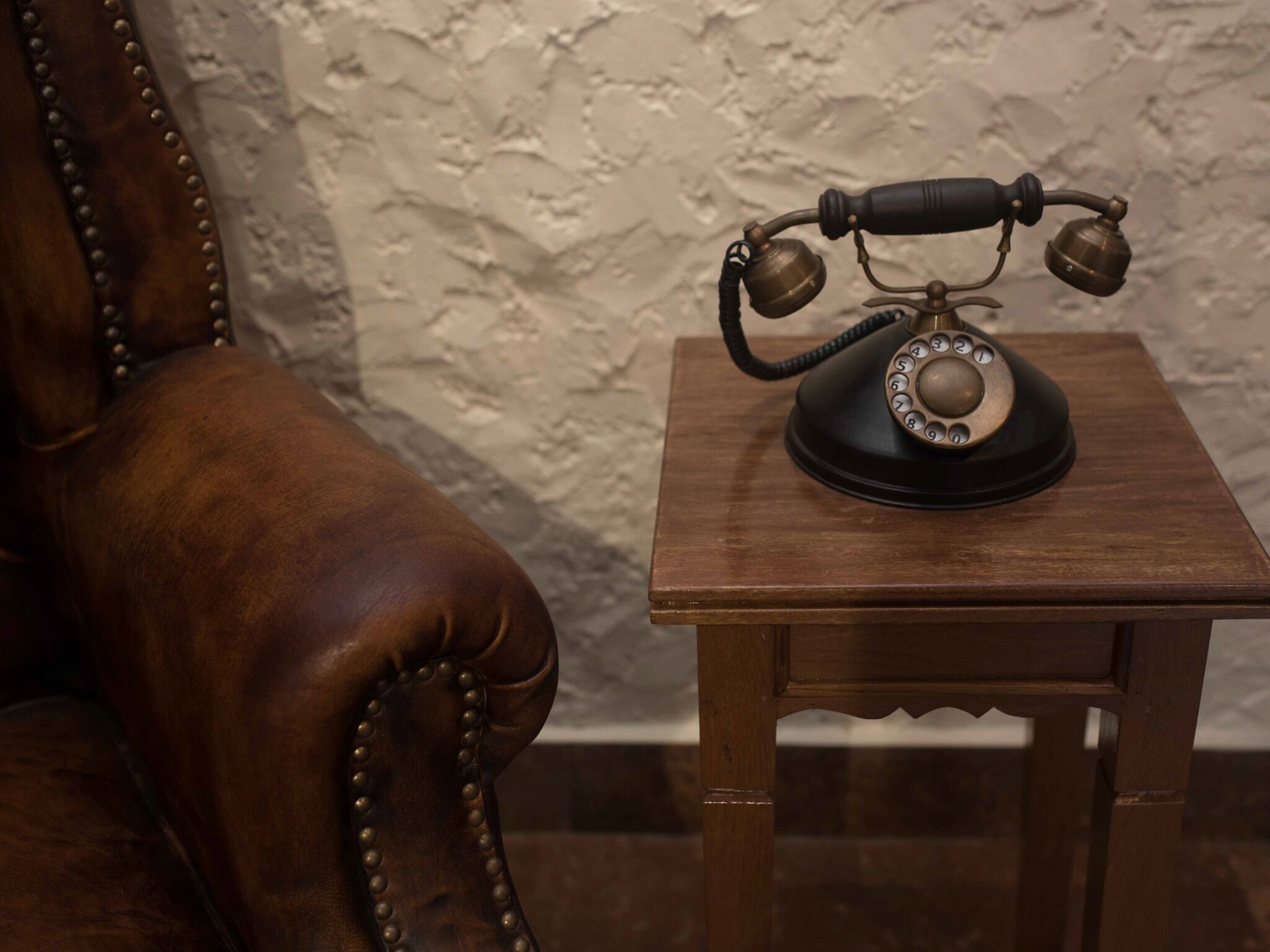 A vintage rotary dial telephone on a wooden table next to a leather armchair.