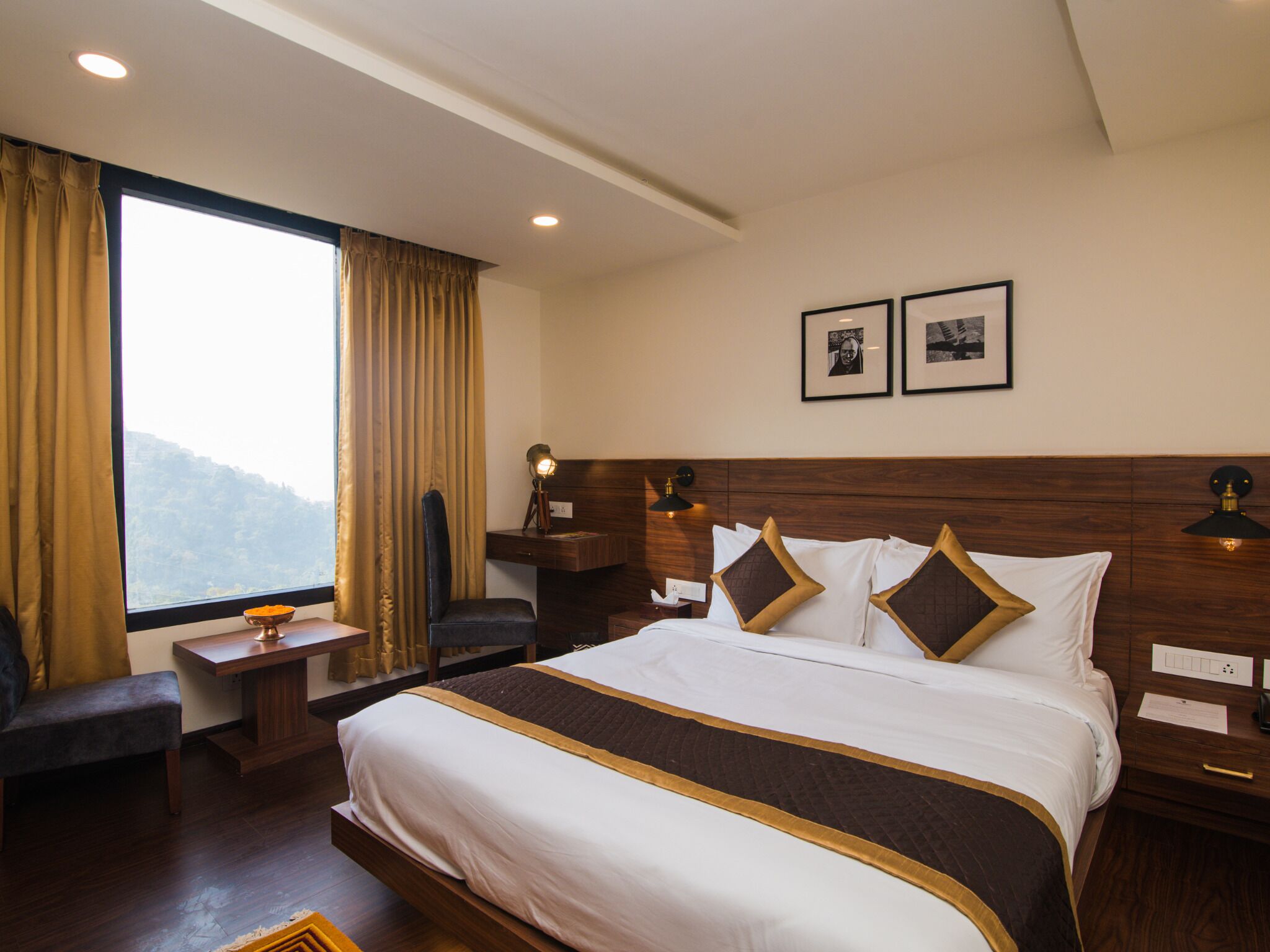 Quarto de hotel com uma cama grande, uma escrivaninha, uma cadeira e uma janela com cortinas. Há duas molduras com imagens na parede.