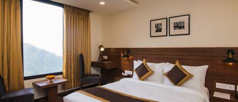 Quarto de hotel com uma cama grande, uma escrivaninha, uma cadeira e uma janela com cortinas. Há duas molduras com imagens na parede.
