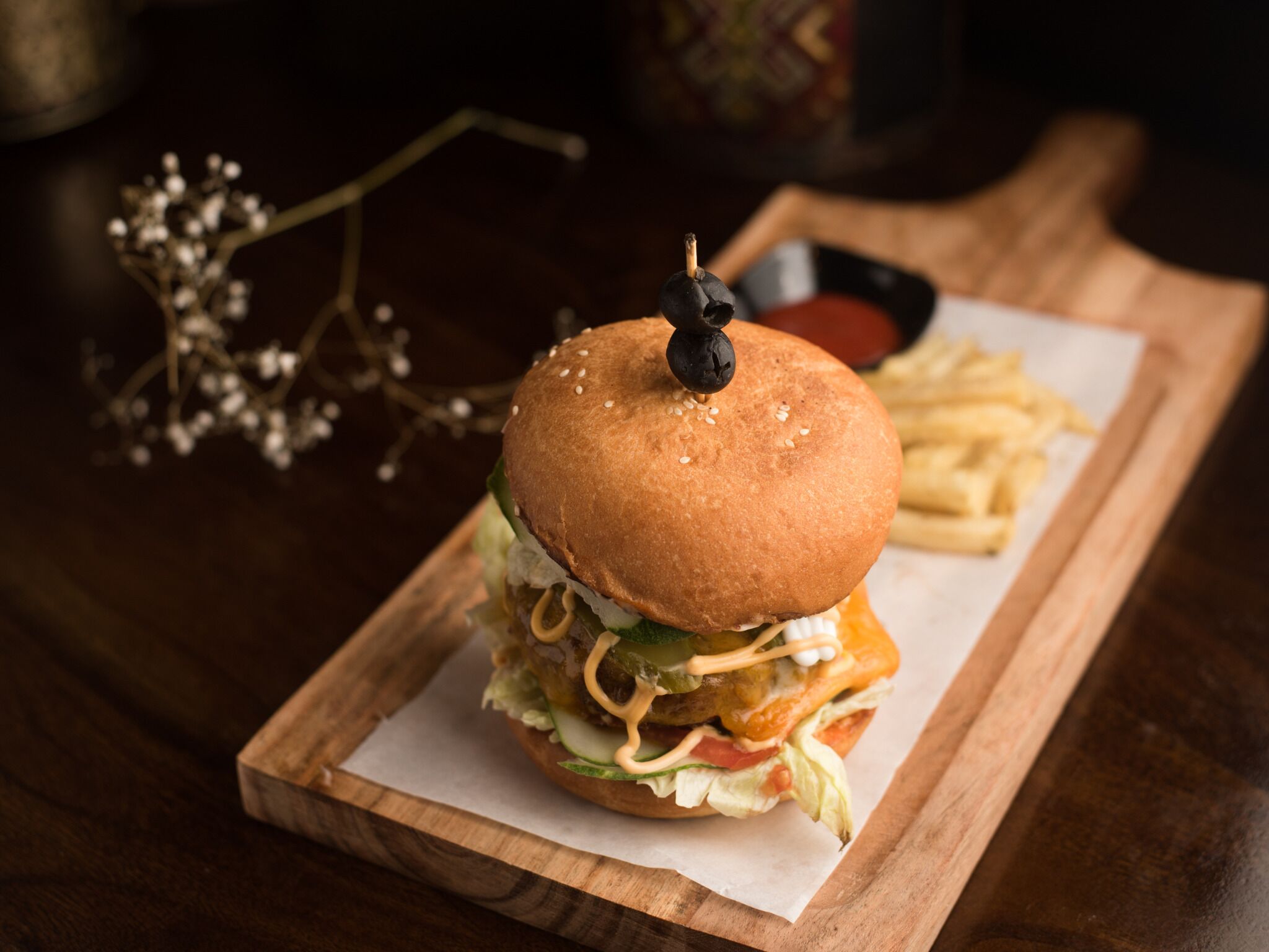 Un hamburger servi dans un pain aux graines de sésame, garni d’oignon mariné, de laitue, de tomate et d’une sauce, le tout présenté sur une planche en bois avec des frites et un petit ramequin de sauce.