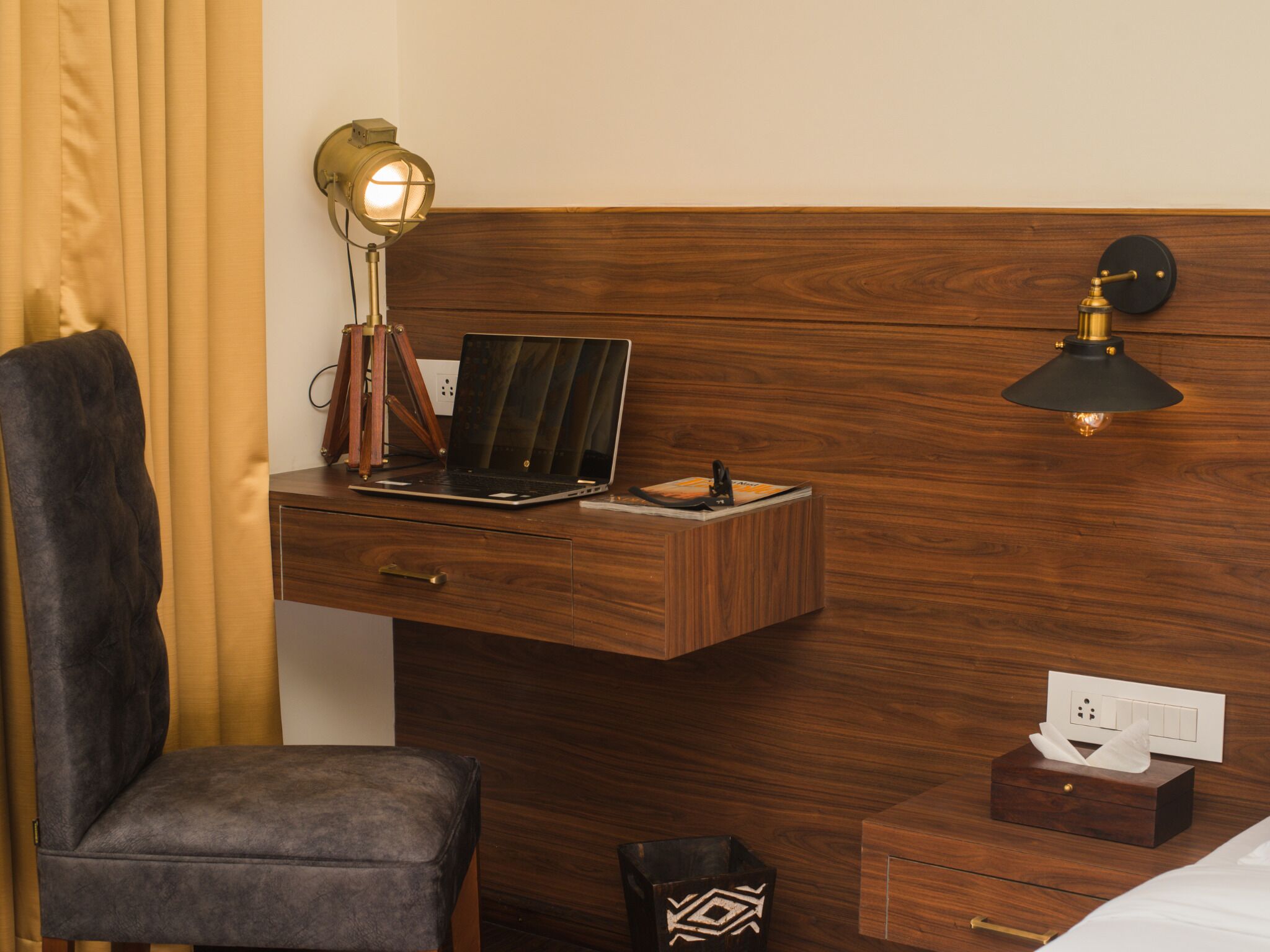 Hotellihuone, jossa on puinen sängynpääty, työpöytä kannettavalla tietokoneella, lamppu, tuoli, yöpöytä paperipyyhekotelolla ja roskakori.