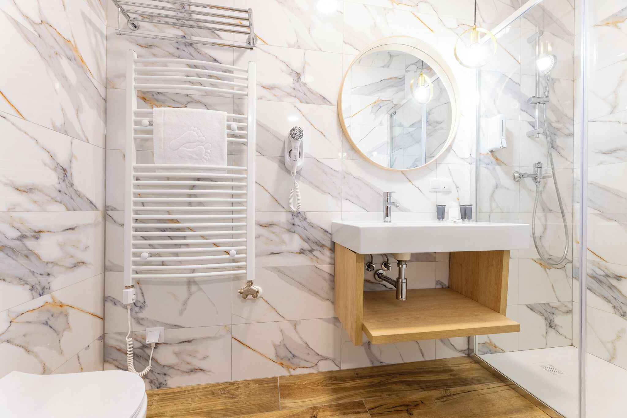 A modern bathroom with a marble floor, a wooden vanity, a white sink, a towel rack, a mirror, and a shower area.