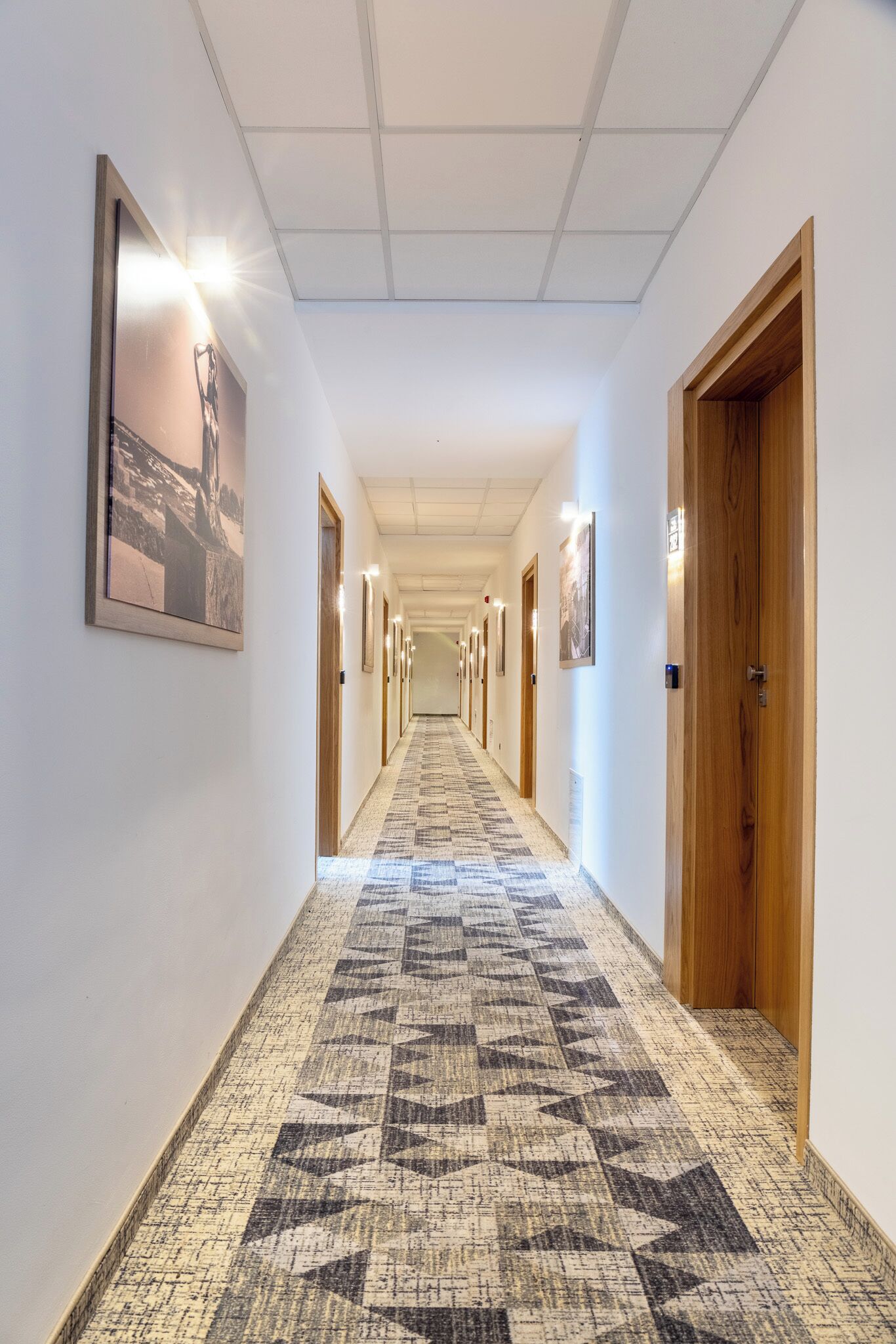 A long, narrow hallway with patterned carpet, wooden doors, and framed pictures on the walls.