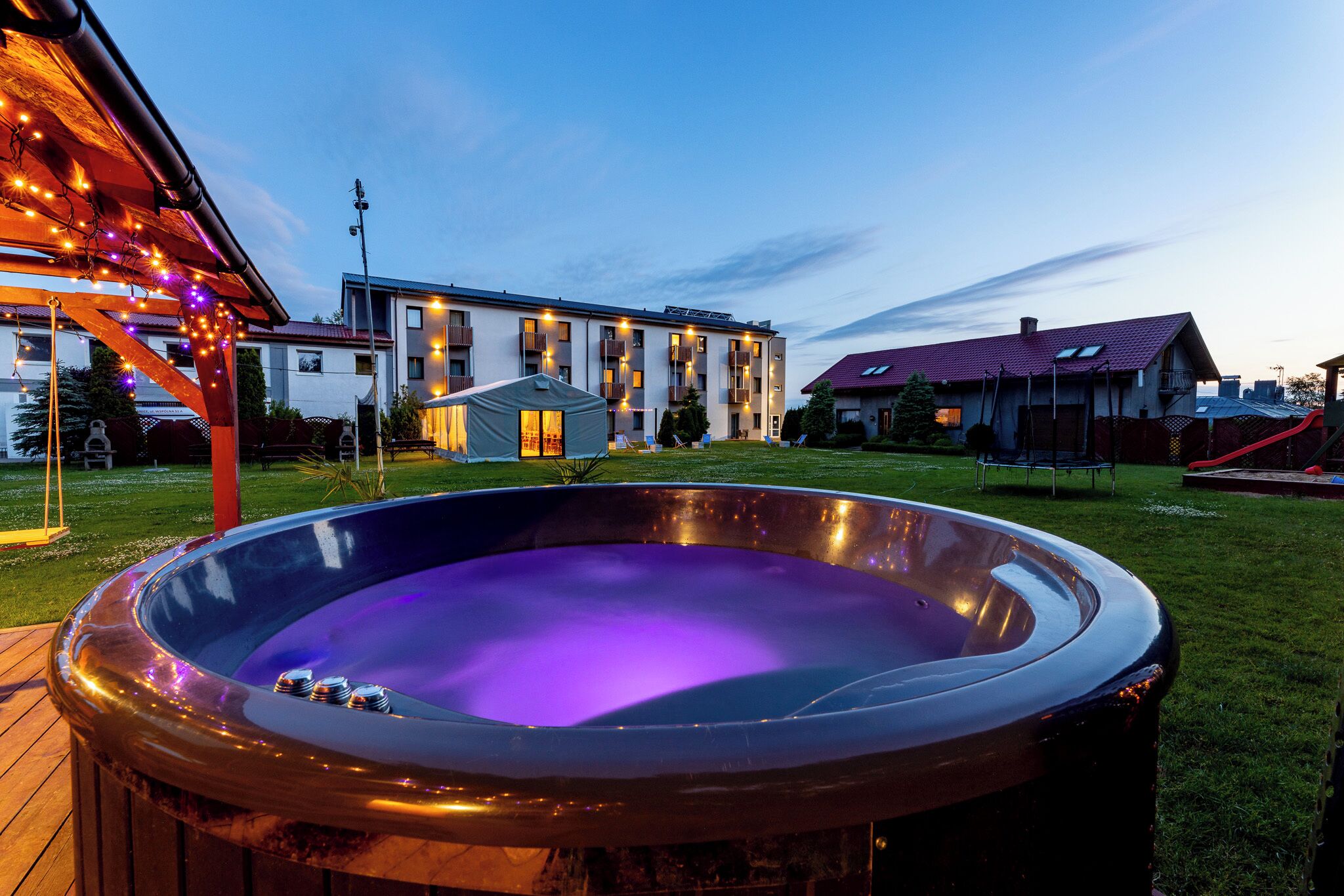Un jacuzzi en un patio trasero con terraza de madera, un porche cubierto con luces colgantes y un edificio moderno al fondo.