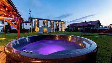Un jacuzzi en un patio trasero con terraza de madera, un porche cubierto con luces colgantes y un edificio moderno al fondo.