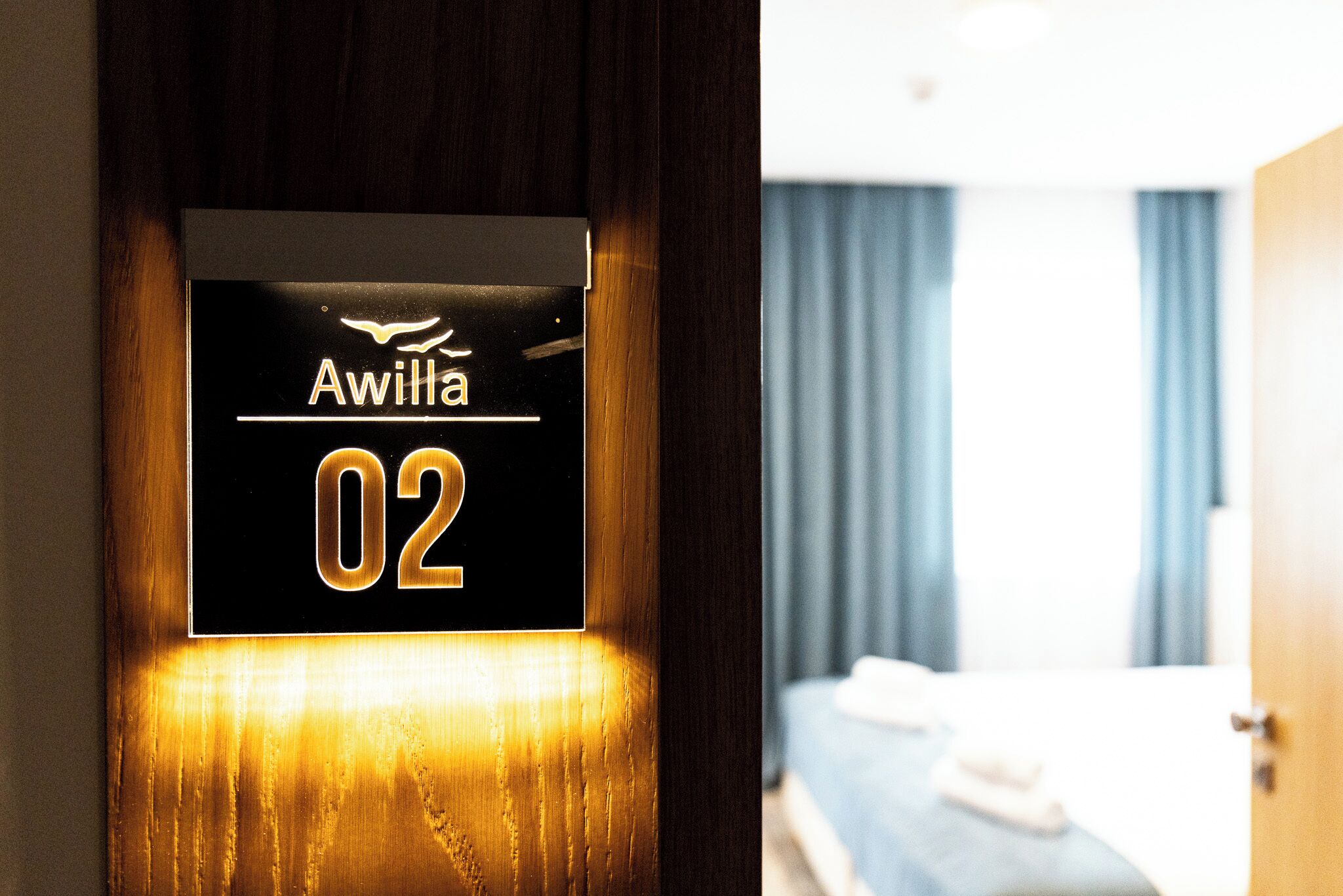 Une porte de chambre d’hôtel avec une pancarte indiquant « Awilla 02 », et un lit recouvert d’une couverture bleue et agrémenté d’oreillers blancs.