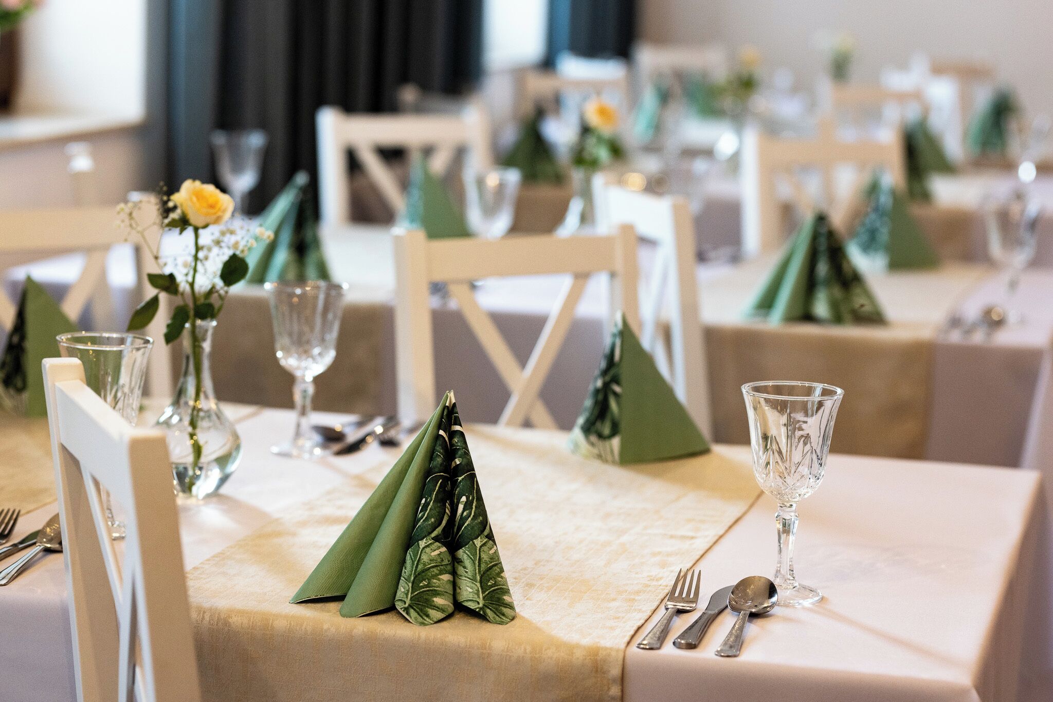 A dining table set with green napkins, clear glassware, and a vase with a yellow flower.