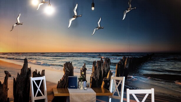 A beachside dining area with a view of the ocean, wooden chairs, a table with a blue menu, and hanging light fixtures. - Awilla (Ustka)