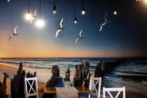 Un área de comedor frente a la playa, con vista al océano, sillas de madera, una mesa con menú azul y lámparas colgantes.