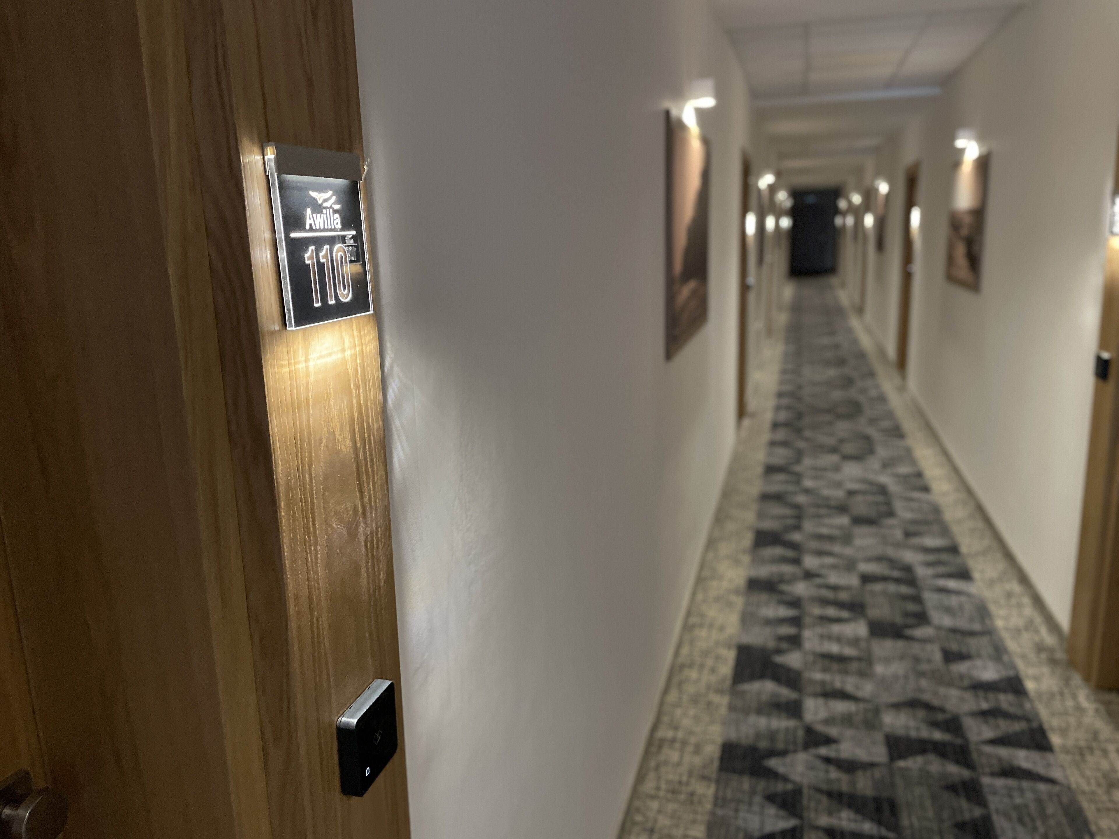 A hallway with a patterned carpet, a door with a sign reading "Awilla 110", and framed pictures on the walls.
