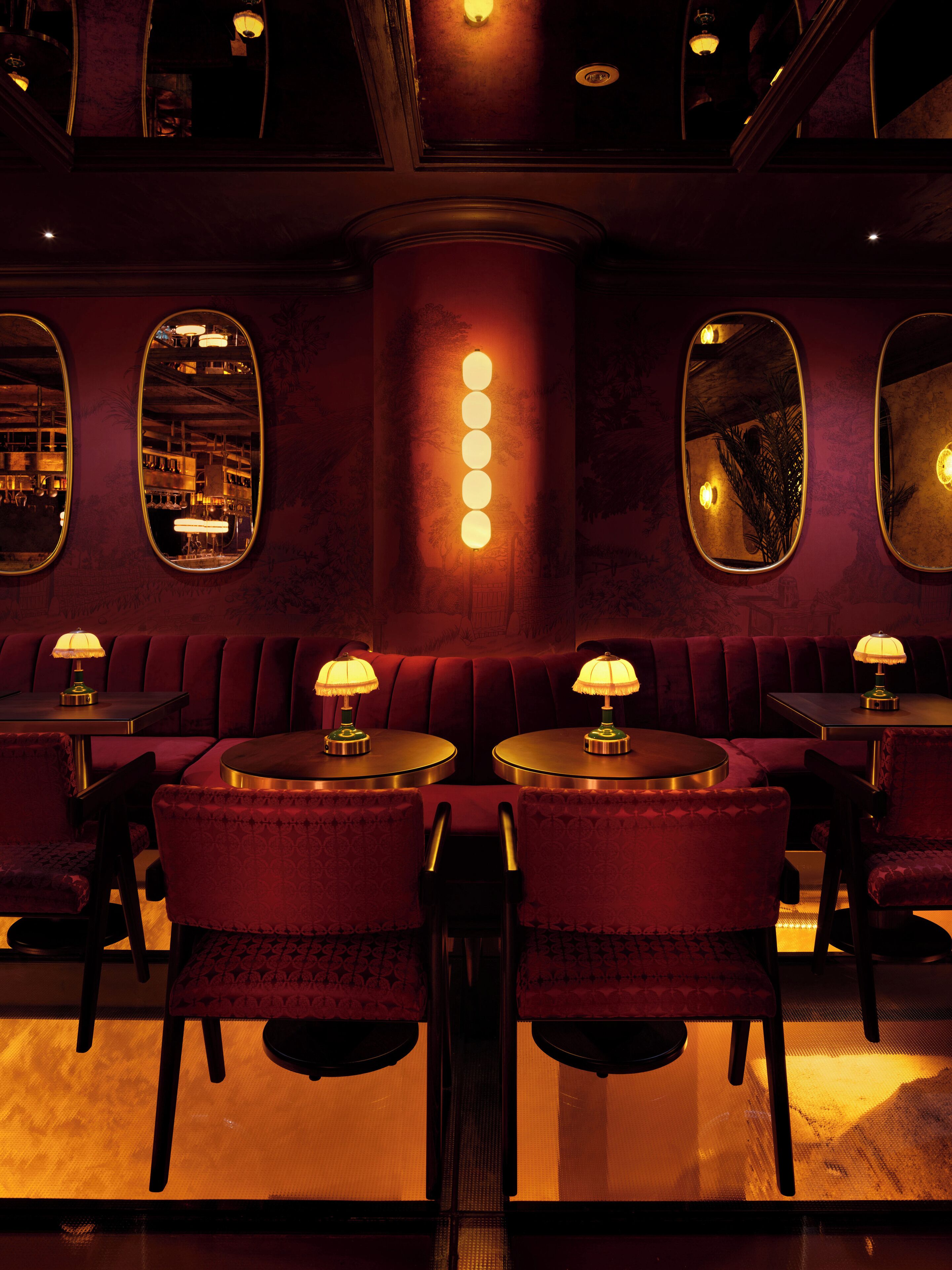 A dimly lit restaurant with red velvet booths and round tables, each with a table lamp.