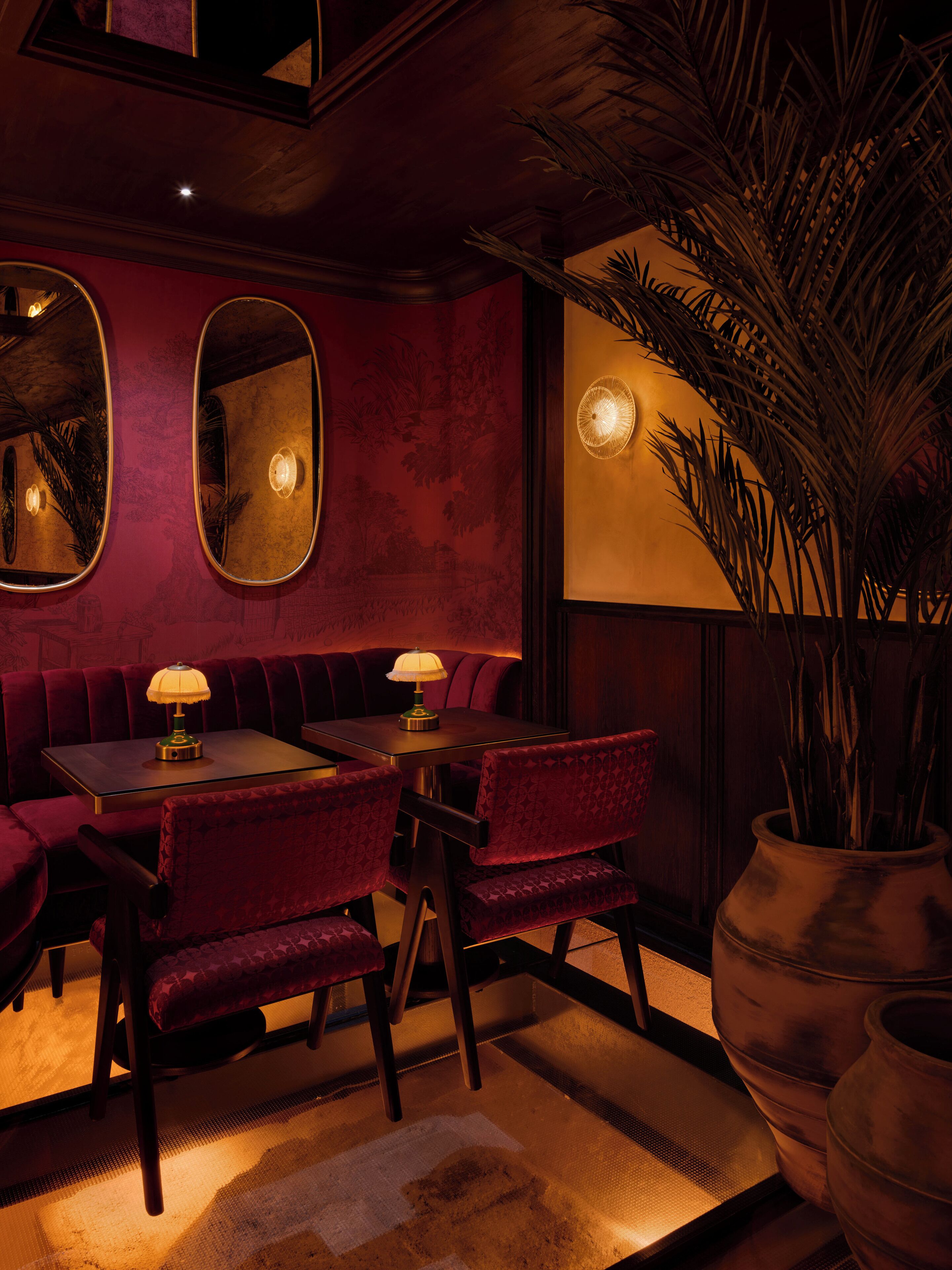 Una habitación tenuemente iluminada con un sofá de terciopelo rojo, una mesa de madera con una lámpara y una planta grande en maceta.