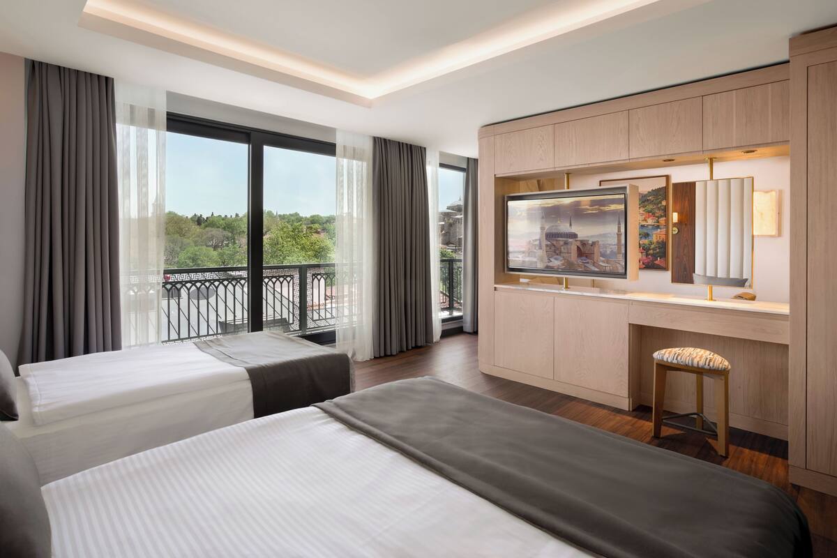 A hotel room with two beds, a large window with a view of greenery, a balcony, and a desk with a TV and a mirror.