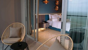 Een moderne hotelkamer met een glazen deur naar een balkon, twee rieten stoelen, een klein rond tafeltje en een bed met een blauwe hoofdeinde.