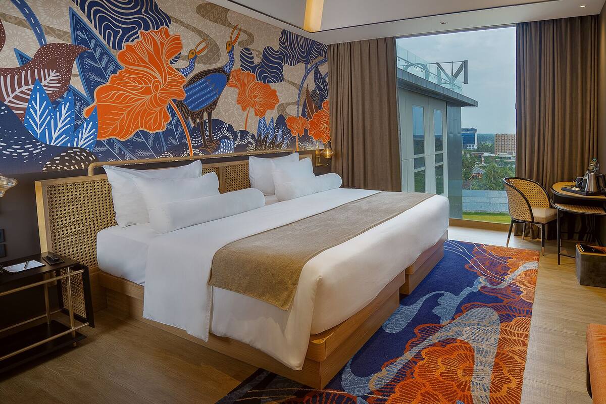 A hotel room with a large bed, a desk with a chair, a view of the city, and a decorative wall mural.