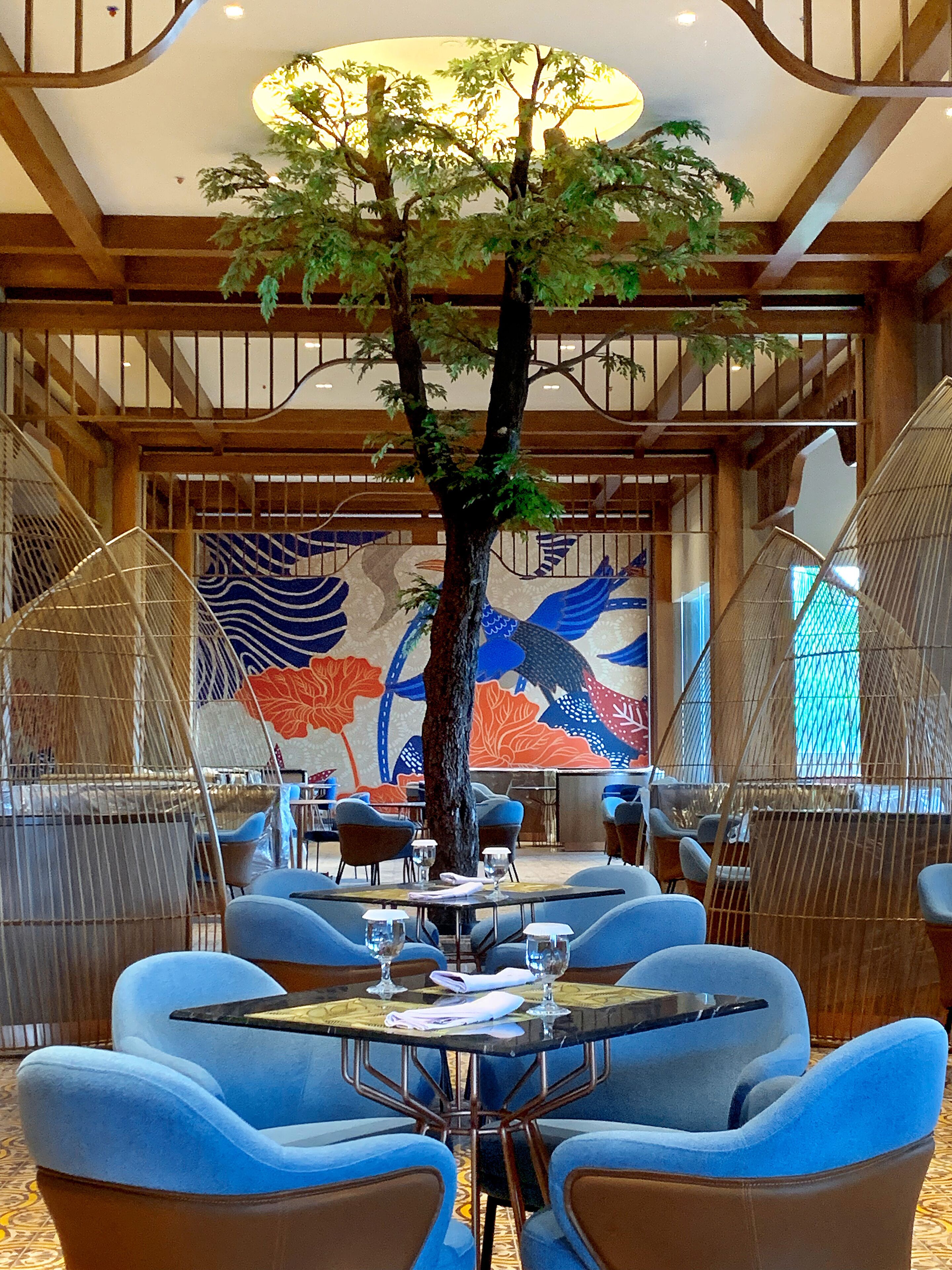 A restaurant with round tables, blue chairs, and a large tree in the center.
