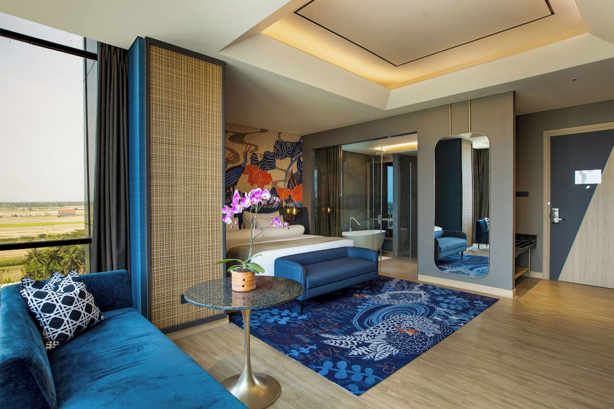 A modern hotel room with a large window, a blue sofa, a small round table, a potted plant, and a bathroom visible through a glass door.