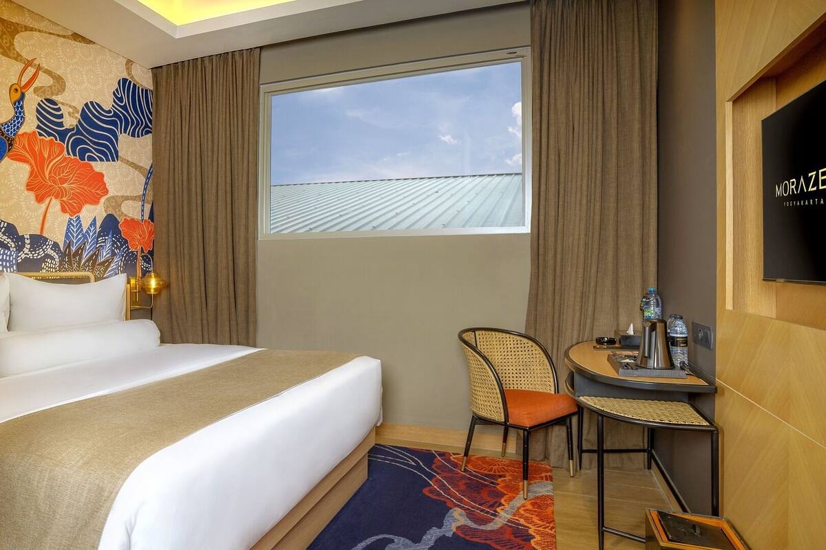 A hotel room with a large bed, a small table with a chair, a window with a view of the roof, and a decorative wall with a bird mural.