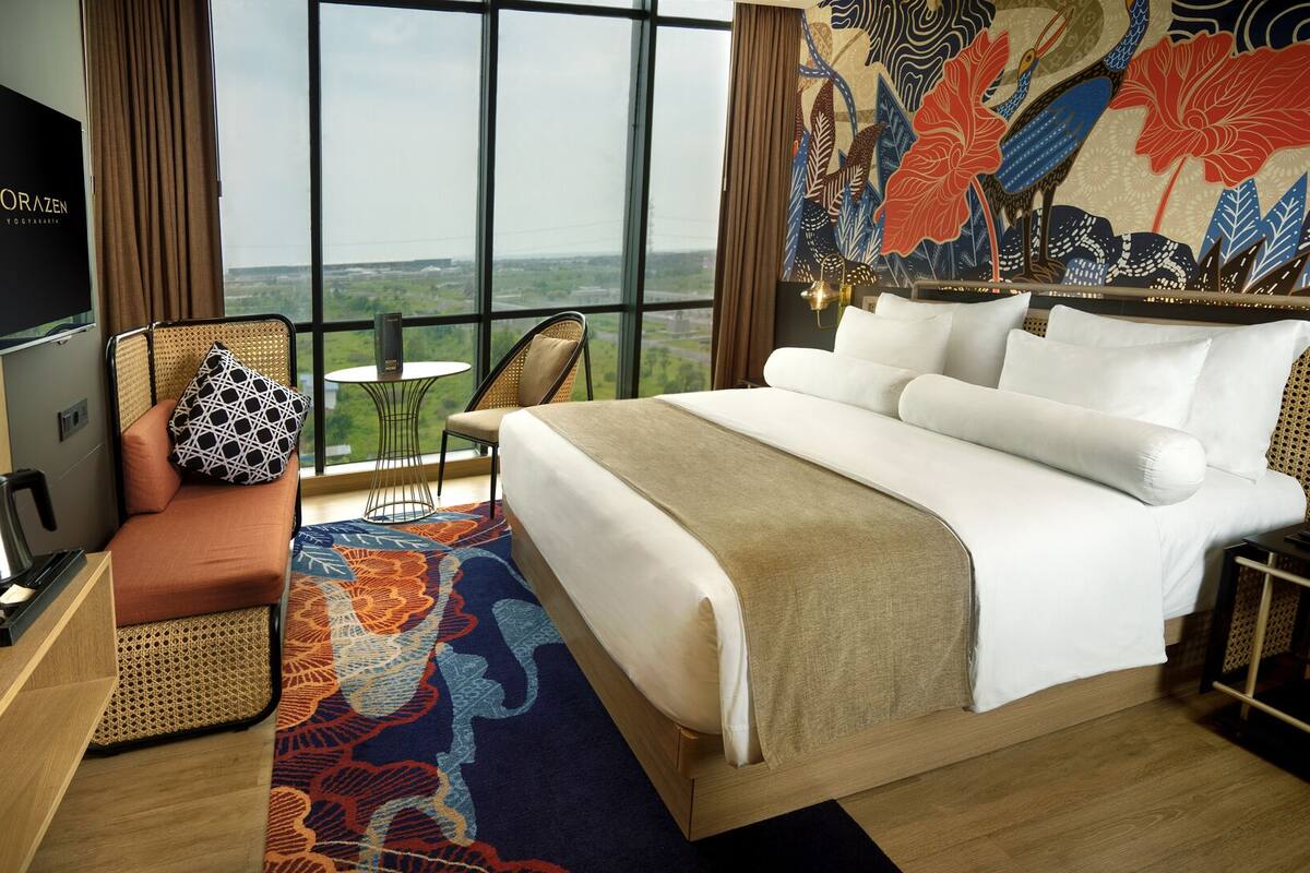 A hotel room with a large bed, a TV, a seating area with a patterned cushion, and a view of the landscape through the window.