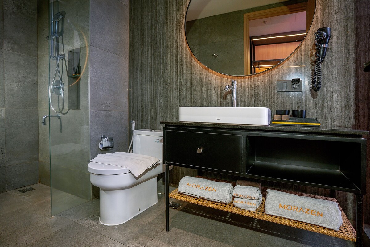 Un baño moderno con ducha envuelta en vidrio, inodoro blanco, tocador negro con espejo y toallas con la marca 'MORAZEN'.