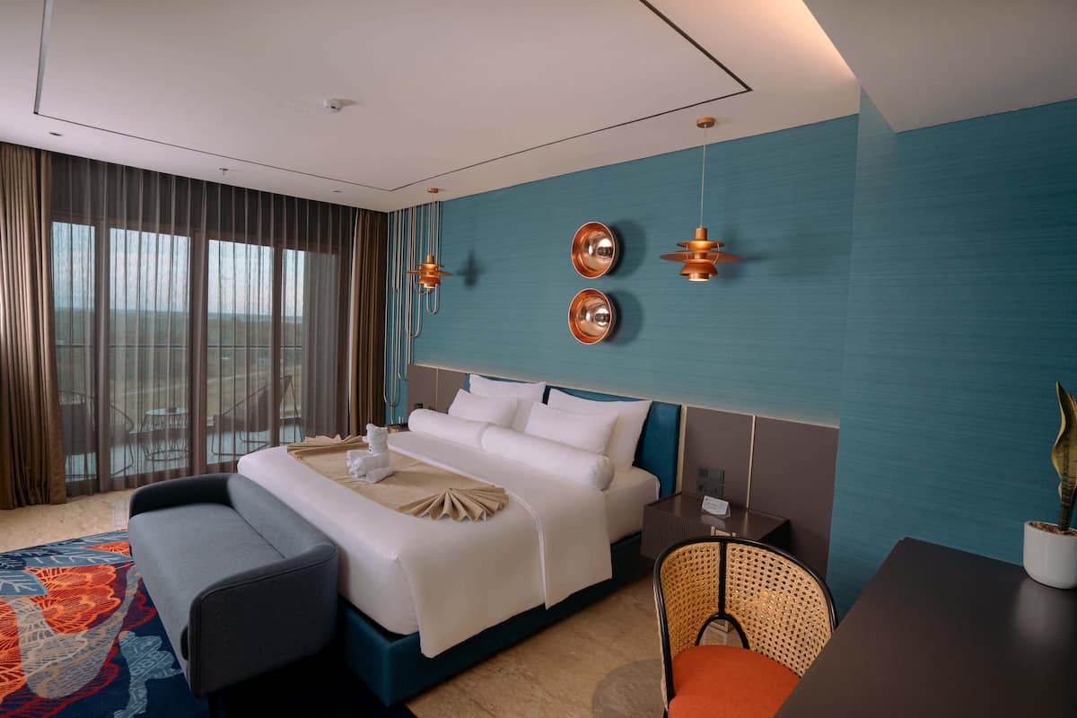 A hotel room with a large bed, a desk, a chair, and a view of the city through large windows.