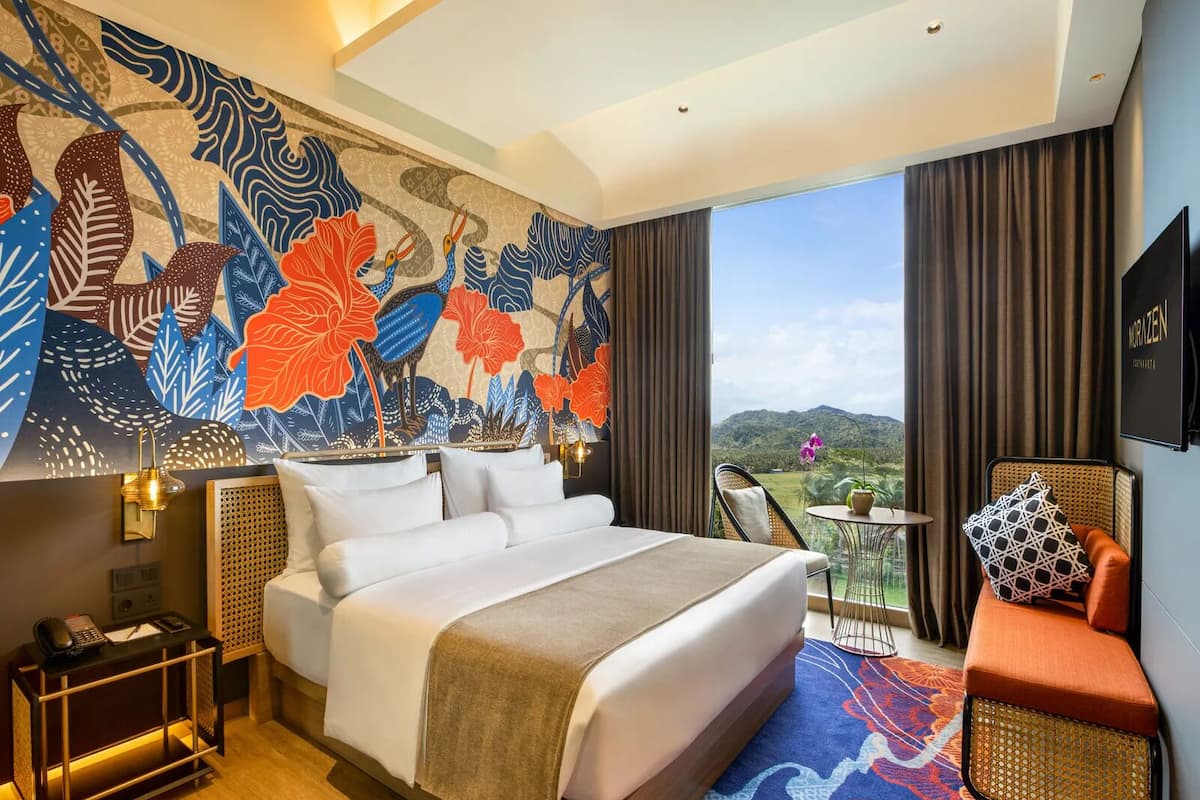 A hotel room with a large bed, a small table, a chair, a sofa, a decorative wall, and a view of the outdoors.