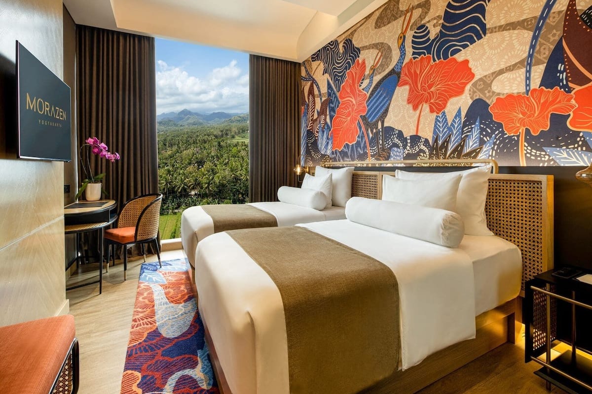 A hotel room with two beds, a desk, a chair, a view of the mountains, and a large floral mural.
