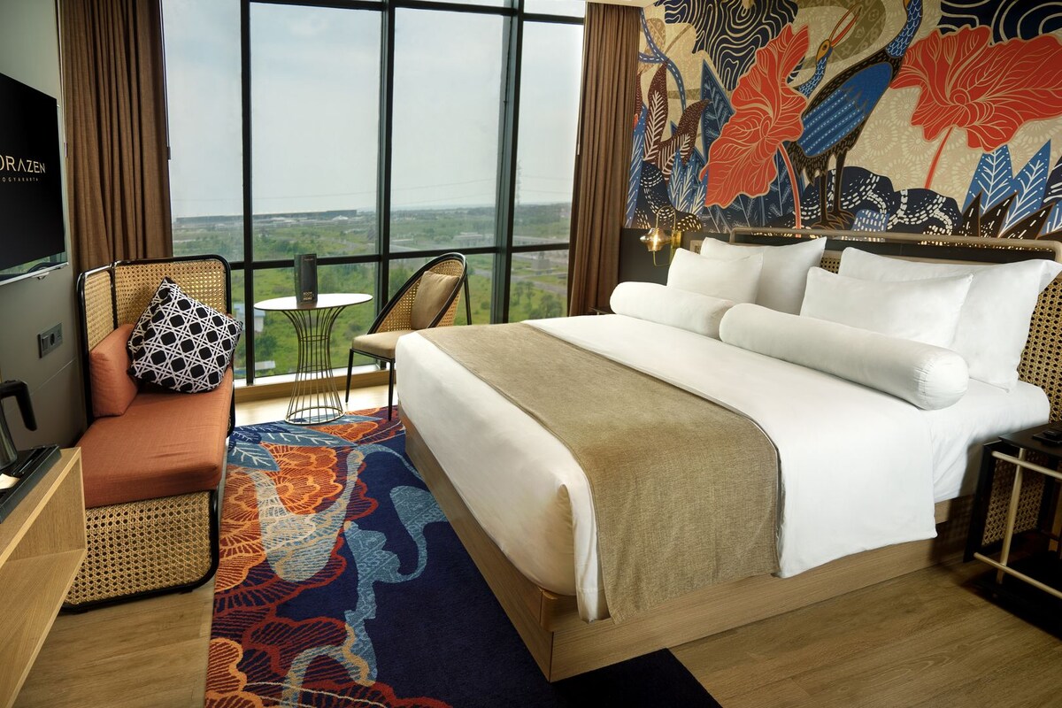 Habitación de hotel con una cama grande, televisión, zona de estar con un cojín estampado y vistas al paisaje por la ventana.