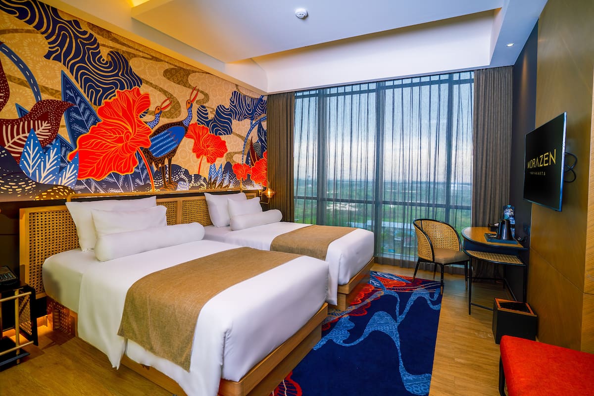 A hotel room with two beds, a desk, a chair, a TV, and a large window with a view.